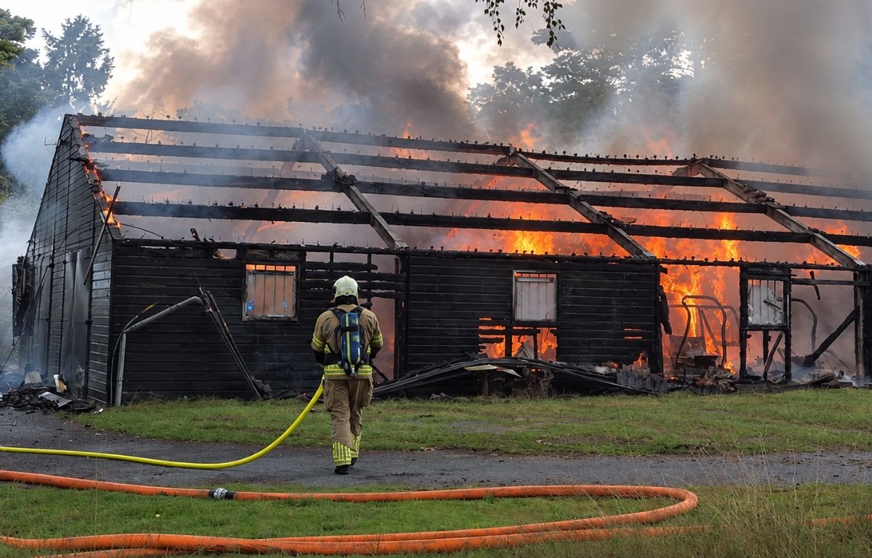 Feuerwehrmann mit einem L&ouml;schschlauch vor einem brennenden Holzschuppen mit Flammen und Rauch